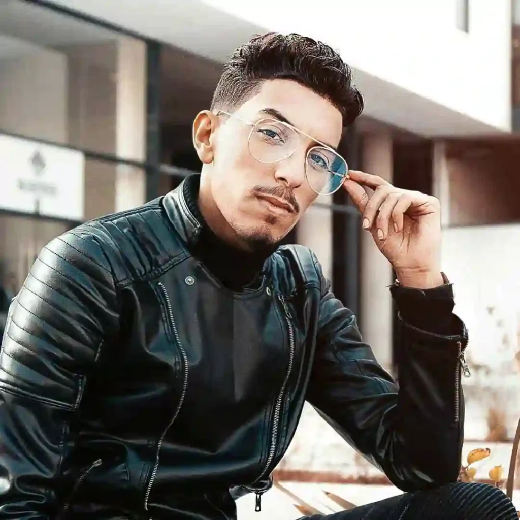 A person in a stylish black leather jacket sits casually, holding a finger to their lips, against a modern architectural backdrop, showcasing an Edgar haircut for men.