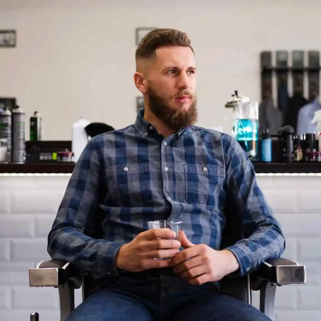 A man seated in a barber chair, wearing a blue plaid shirt, holding a glass in a modern barbershop setting, showcasing Edgar haircuts for men.