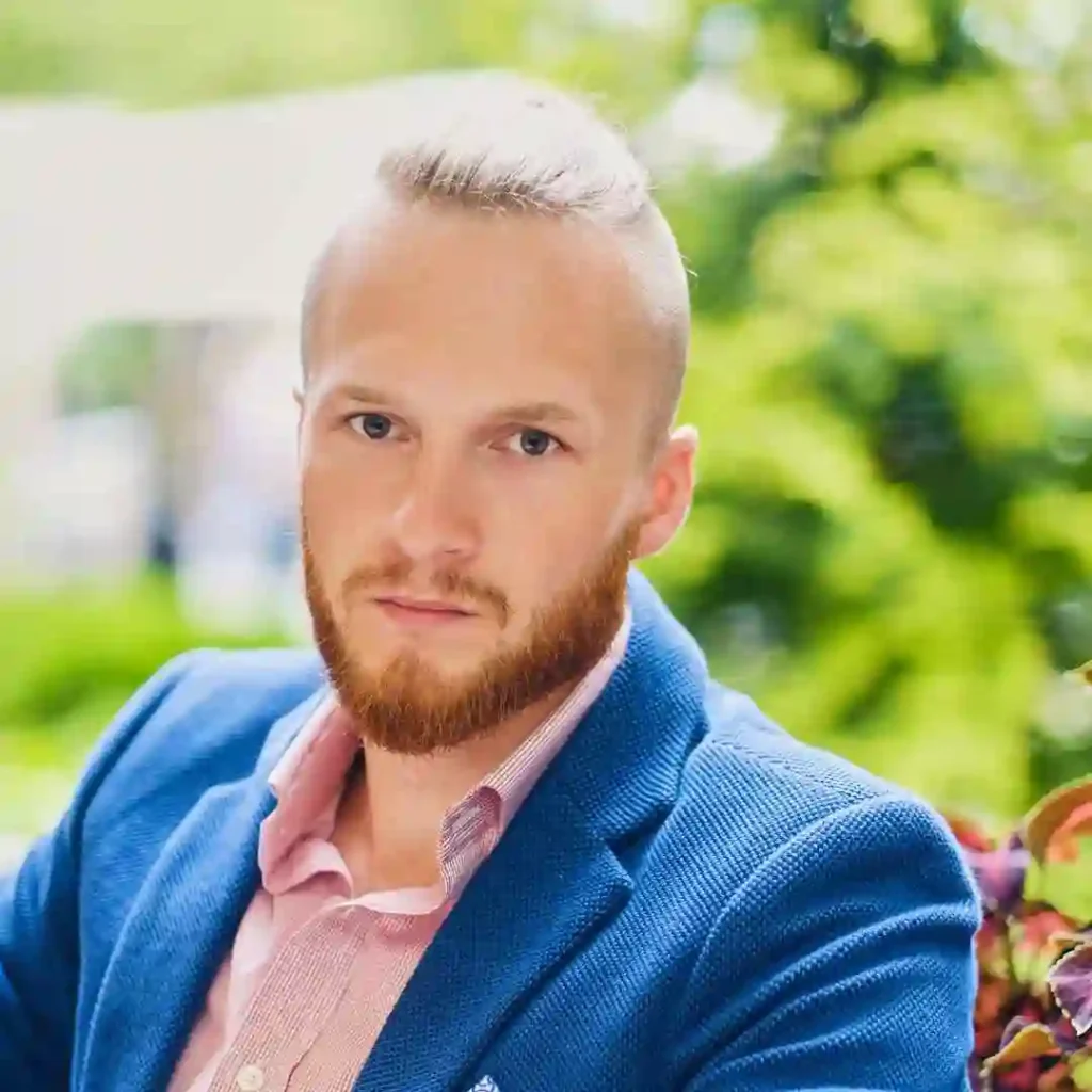 A man with Edgar haircuts for men, wearing a blue blazer and pink shirt, relaxes outdoors, surrounded by green foliage and colorful plants.