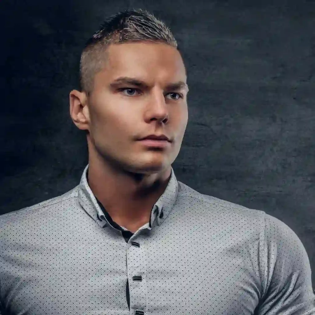 A man wearing a light gray button-up shirt with a subtle pattern stands against a dark textured background, showcasing an Edgar haircut for men.