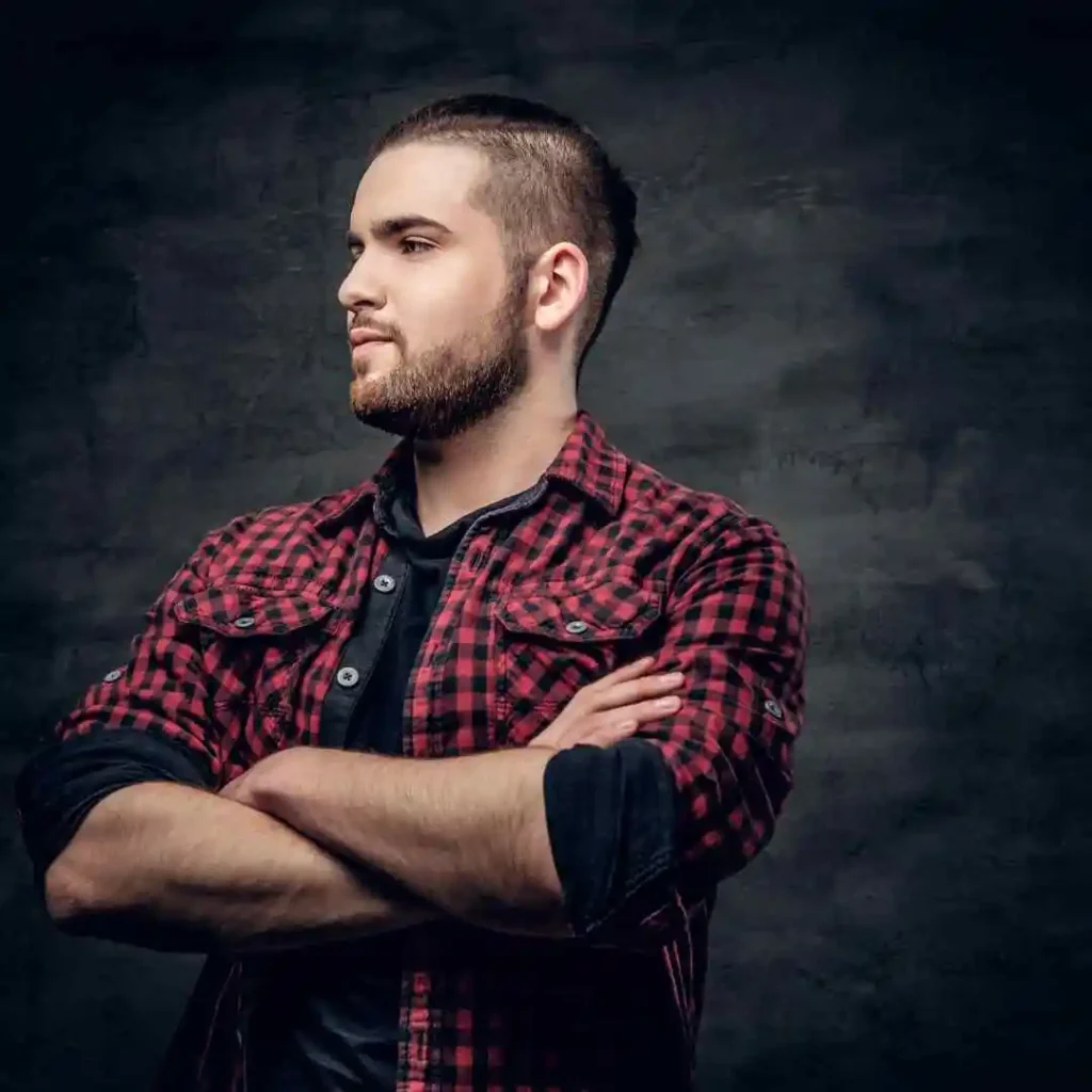 A man in a red and black plaid shirt stands confidently with arms crossed, showcasing an Edgar haircut for men, set against a dark, textured background.