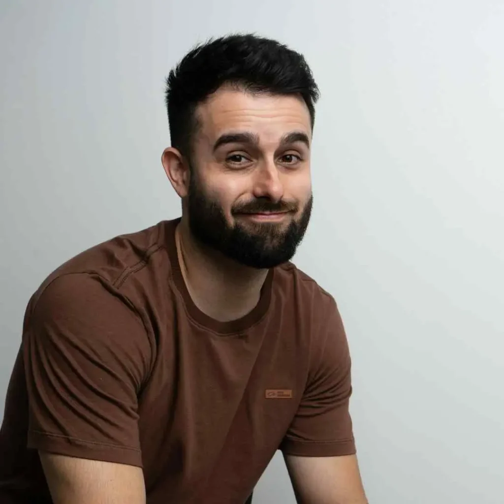 A young man with dark hair wearing a brown t-shirt, seated against a light gray background, showcasing an Edgar haircut for men, creating a minimalist portrait.