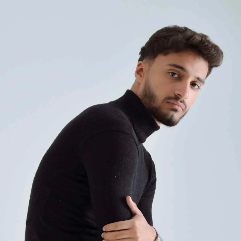 A person wearing a black turtleneck sweater stands in profile, showing off an Edgar haircut for men, with their arms crossed against a light gray background.