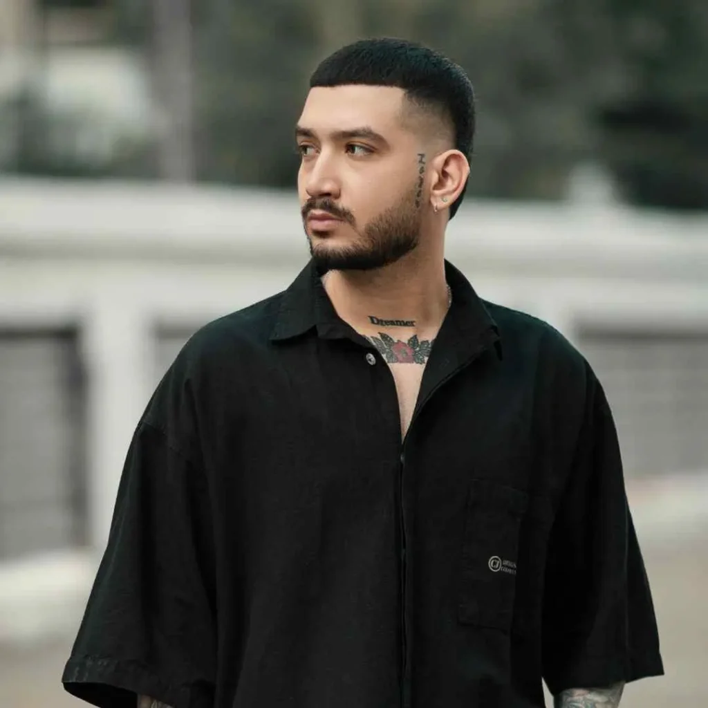 Edgar Hairstyles for Men- Top Styles You Needs to Try 6 A person with an Edgar haircut for men, wearing a short-sleeved black shirt, showcases various tattoos on their chest and neck, set against an outdoor background.