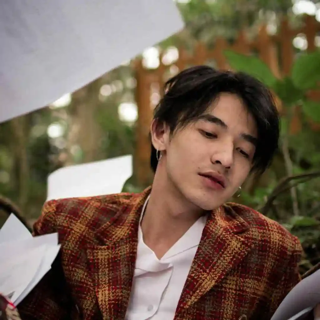 A person showcasing MEN HAIRSTYLES CURTAINS, dressed in a textured, brown plaid jacket, is surrounded by scattered papers in a lush, green outdoor setting.