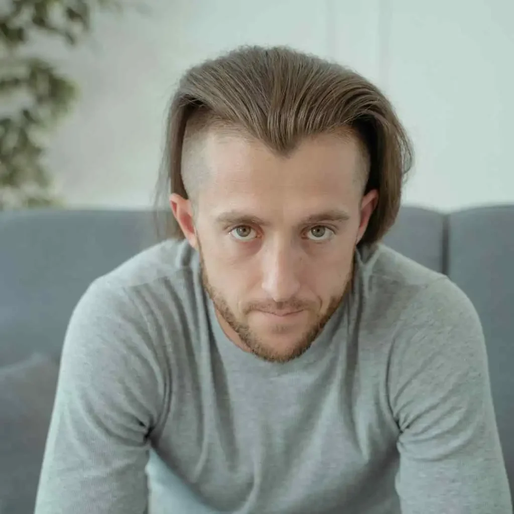 A person with long, styled hair showcasing MEN HAIRSTYLES CURTAINS sits on a grey couch, wearing a light grey sweater, against a soft, blurred background with greenery.