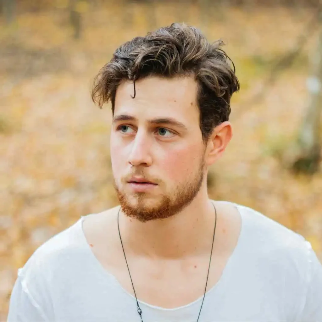 A young man with Edgar haircuts for men and tousled hair wears a white shirt and a necklace, standing in a forest with autumn leaves scattered around him.