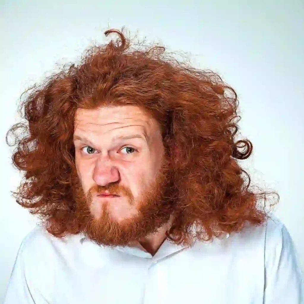 A person with vibrant red, curly hair wearing a white shirt, showcasing an Edgar haircut for men, set against a light, neutral background.
