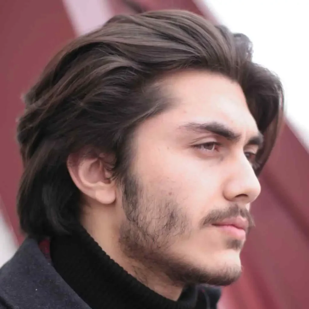 A person with long, dark hair and wearing a black turtleneck stands against a blurred red background, exuding a casual vibe, showcasing men hairstyles curtains.