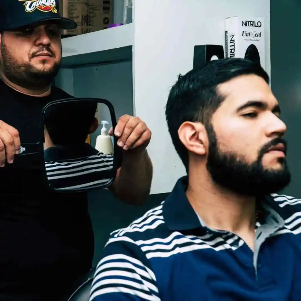 Edgar Hairstyles for Men- Top Styles You Needs to Try 2 A barber holds a mirror to show a customer his Edgar haircut for men, while another person sits in a chair in a modern barbershop.