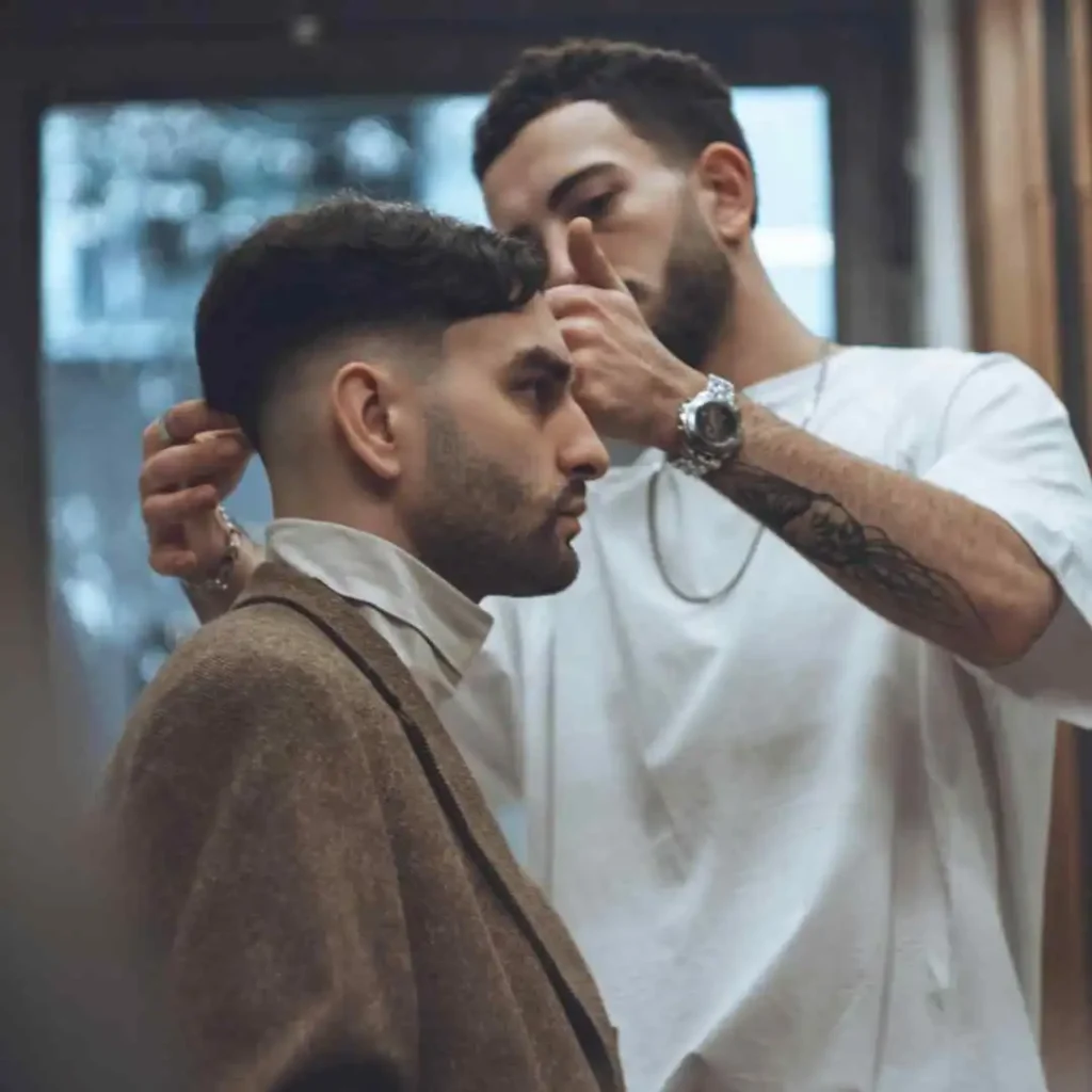 A barber styles a client's hair in a cozy salon, highlighting an elegant brown suit and a white shirt layered underneath, featuring Edgar haircuts for men.