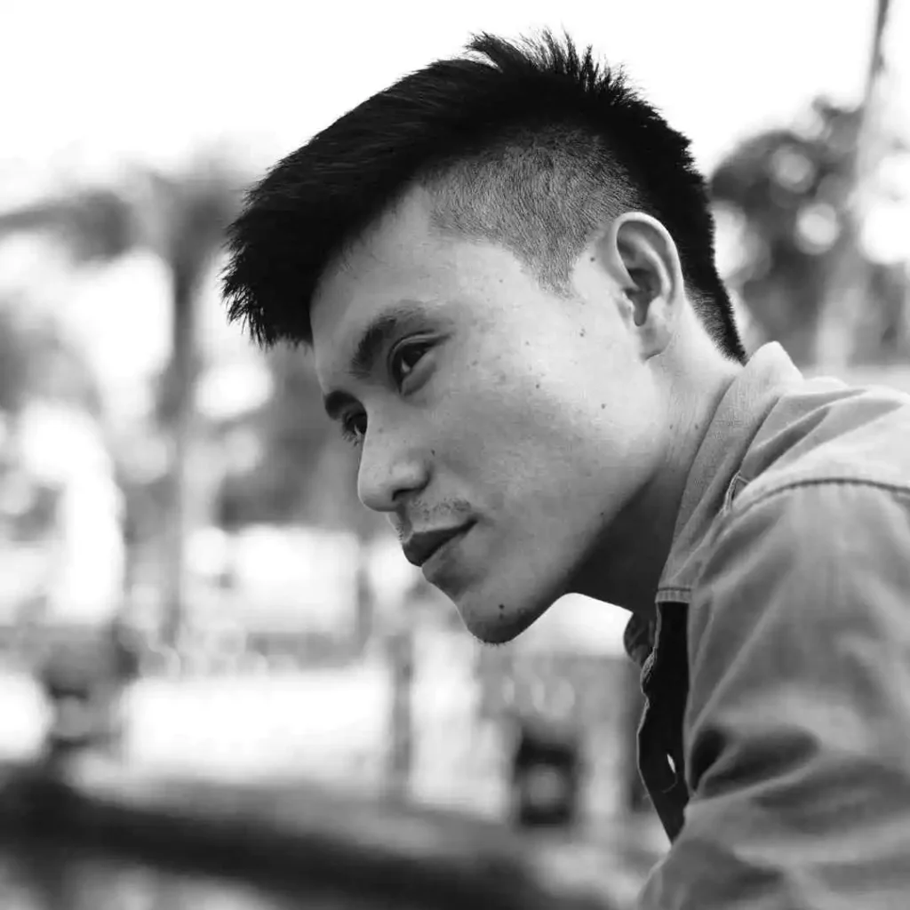 "A young person in a denim jacket sits pensively outdoors, showcasing an Edgar haircut for men, with blurred trees in the background creating a tranquil atmosphere.
