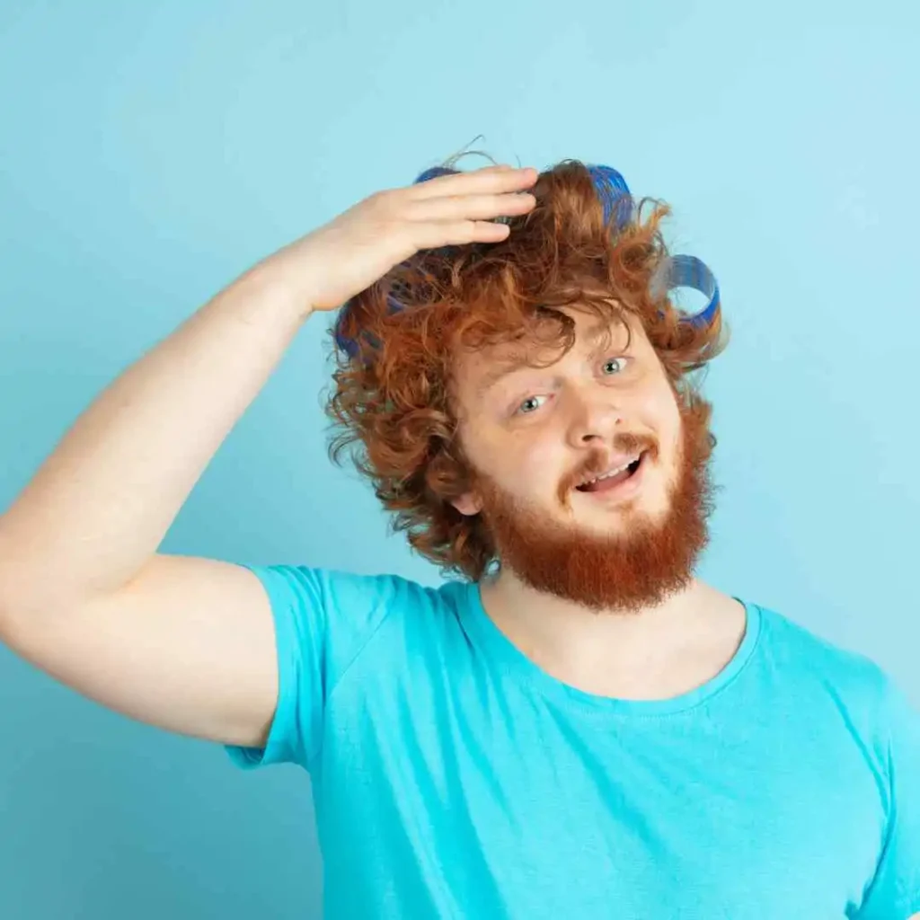 A person with curly red hair and a beard gestures towards their hair while wearing a blue t-shirt, showcasing Edgar haircuts for men against a light blue background.