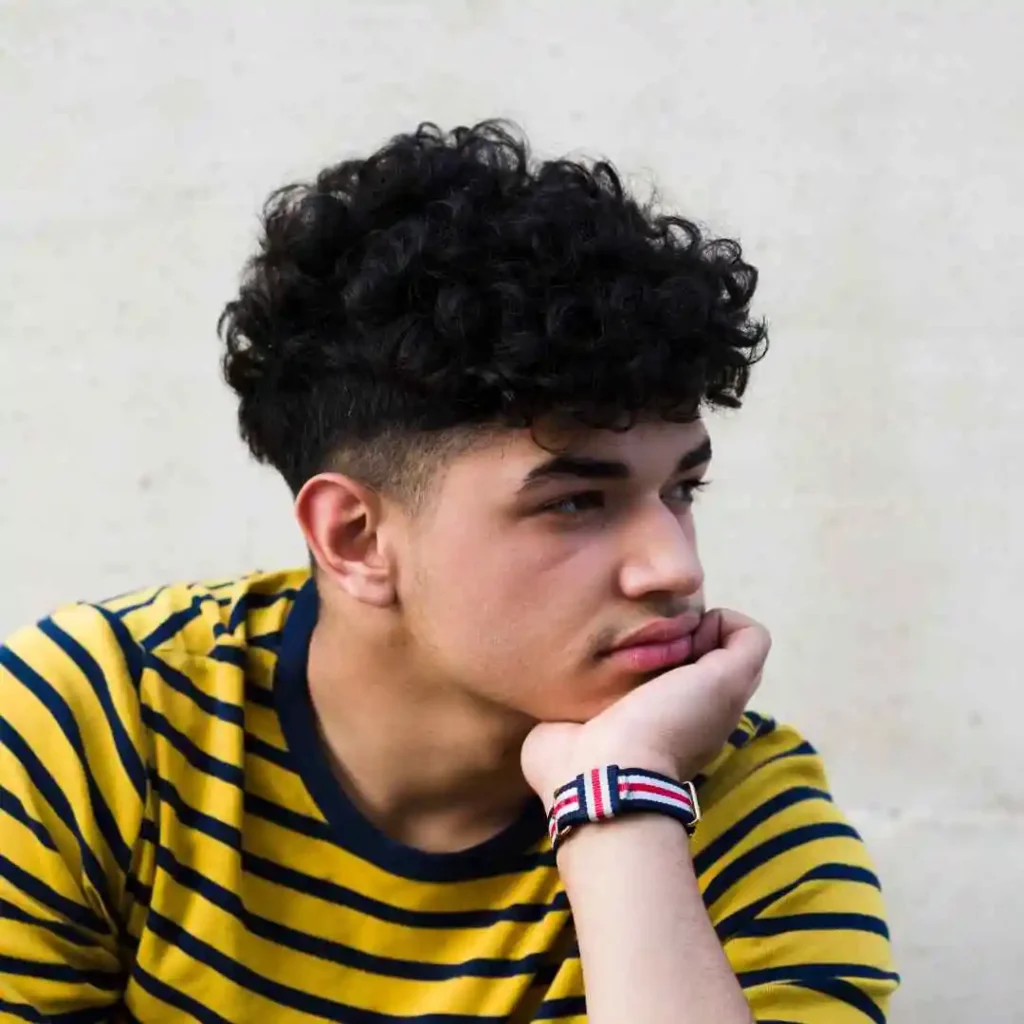 A person with curly hair wearing a yellow and navy striped shirt, resting their chin on their hand against a light gray background, showcasing an Edgar haircut for men.