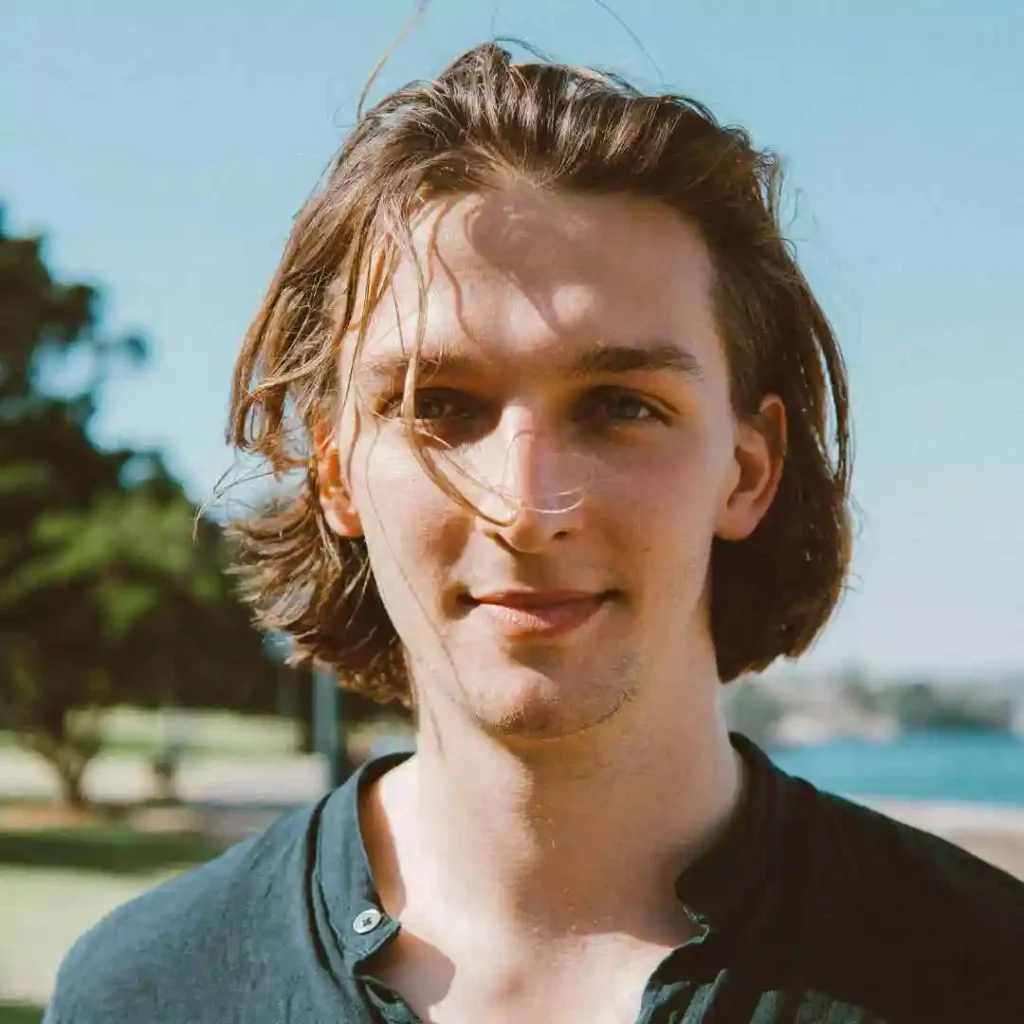 A person with wavy hair styled in men hairstyles curtains, wearing a black button-up shirt, stands outdoors against a clear blue sky and scenic backdrop.