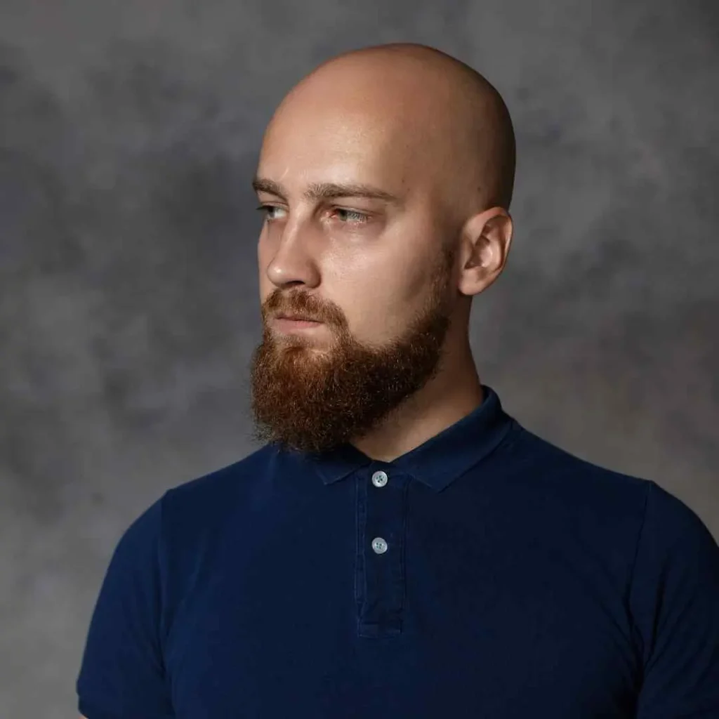 Cool And Breezy Summer Hairstyles for Men 1 A person with a bald head and beard, showcasing one of the trendy summer hairstyles for men, wears a dark blue polo shirt while standing against a textured gray background.