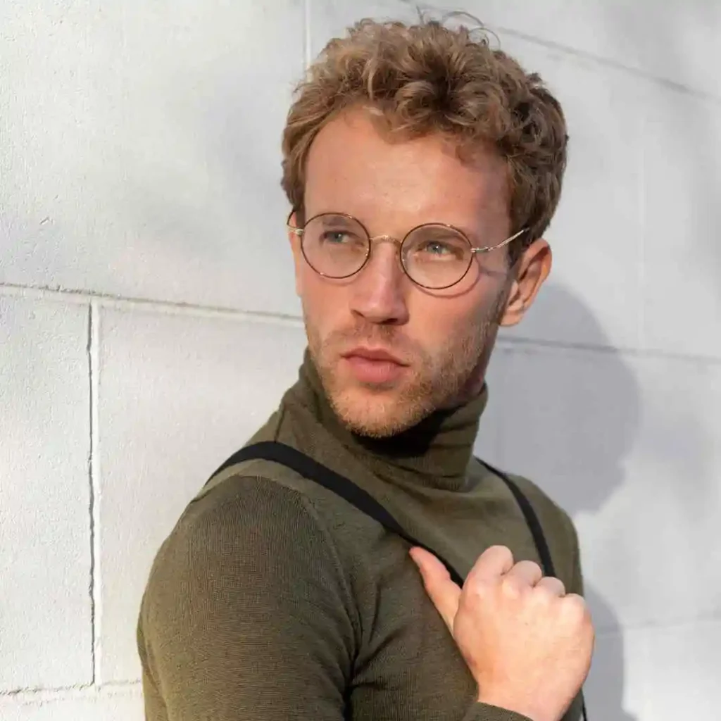 A person showcasing trendy men hairstyles with glasses, wearing a green turtleneck and black suspenders, stands against a light-gray wall, casting a soft shadow.