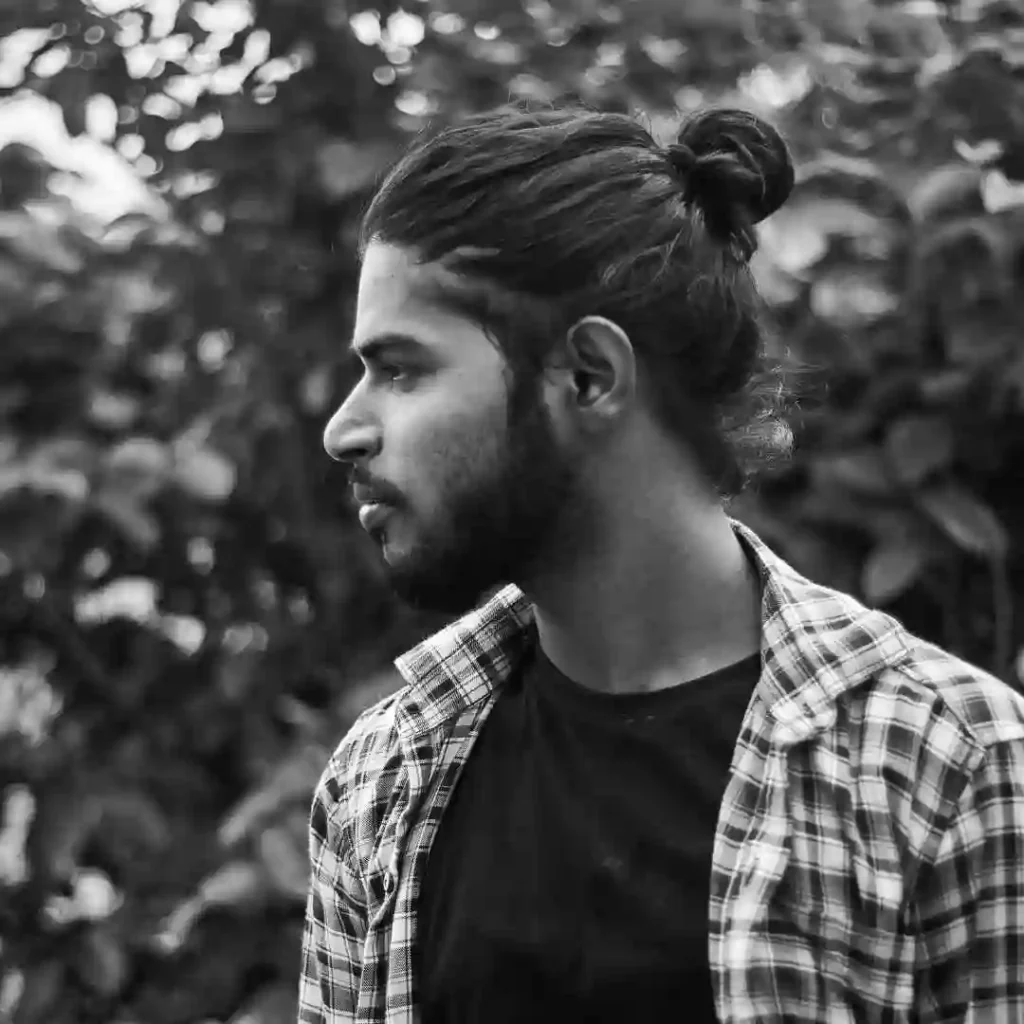 Long Hair Styling for Men.. Creative Hairstyles And Ideas 1 A person with long hair styling for men stands outdoors, wearing a black t-shirt and a checked shirt, against a backdrop of lush greenery in black and white.