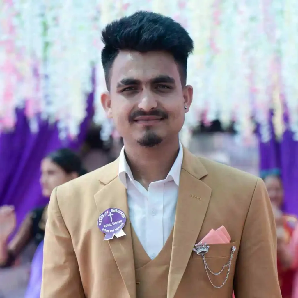 A young man in a tan suit stands confidently, showcasing a stylish wedding hairstyle for men, adorned with a badge and pink accents, against a festive backdrop of colorful decorations.