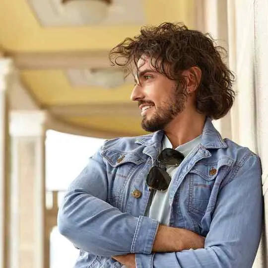 A casually dressed individual stands with arms crossed, wearing a denim jacket and sunglasses, against a softly lit architectural backdrop.