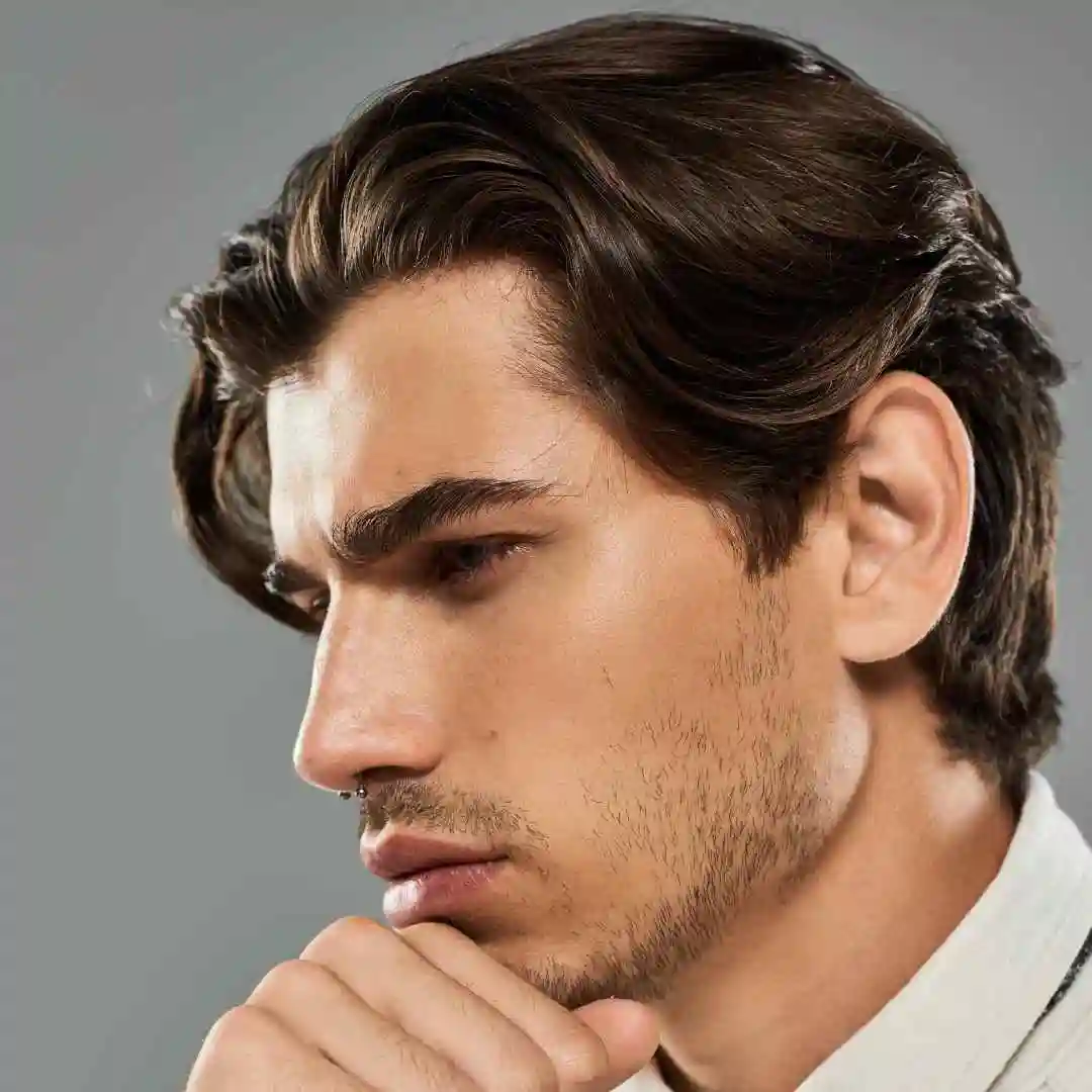 A stylish man in a white shirt poses thoughtfully, showcasing textured, dark hair and a well-groomed ear against a grey background.