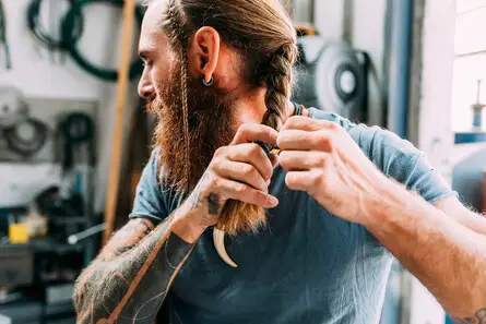 A person with long hair and tattoos is carefully braiding their hair, showcasing one of the trendy summer hairstyles for men, set against a backdrop of workshop tools and equipment.