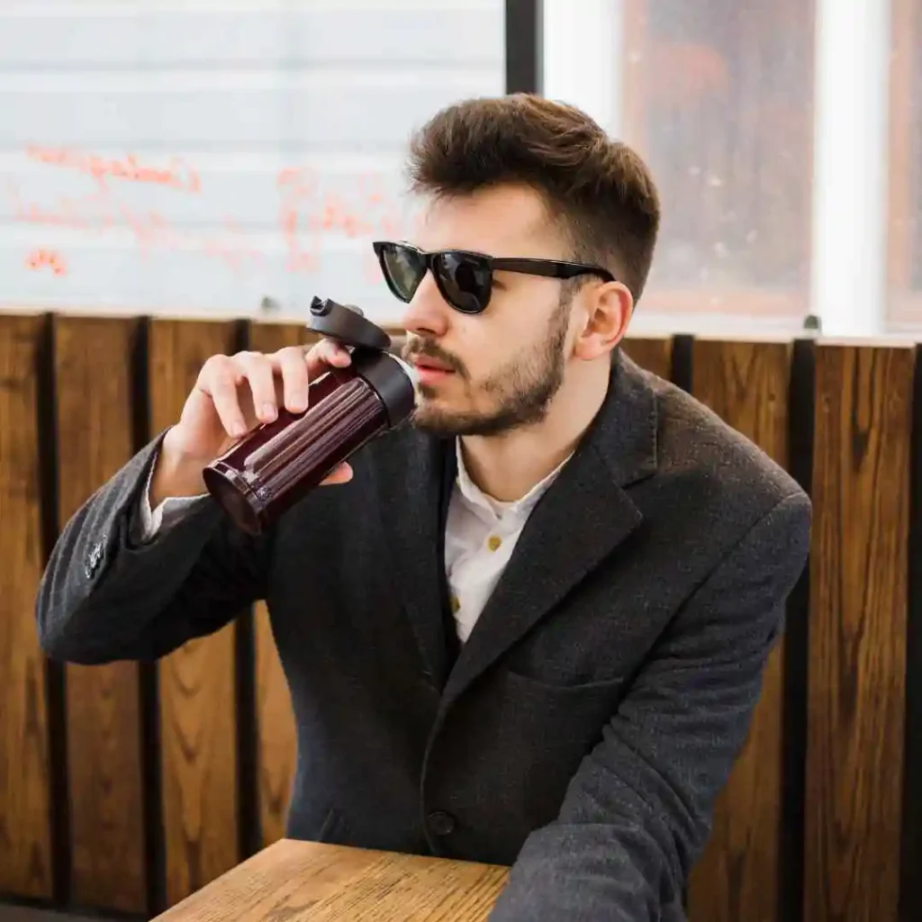 A person with trendy men hairstyles with glasses, dressed in a stylish gray blazer, sips from a red water bottle while seated at a wooden table in a cozy café setting.