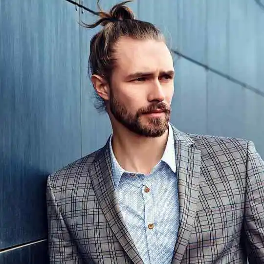 A stylish man with long hair in a bun wears a plaid suit jacket over a light blue shirt, posing against a modern blue wall, showcasing a wedding hairstyle for men.