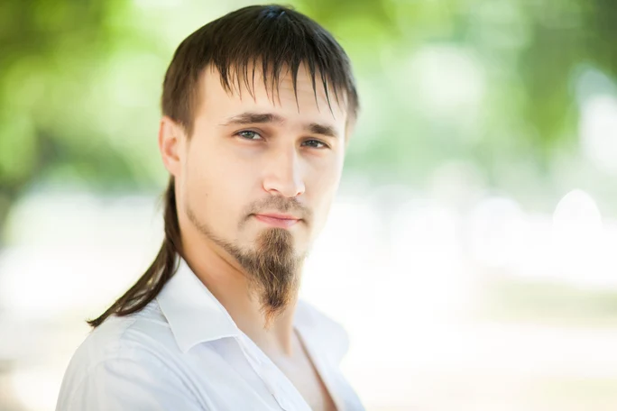 A person with dark hair styled in a ponytail wears a white shirt, posed outdoors with a soft-focus green background, showcasing a FADE HAIRSTYLE FOR MEN.