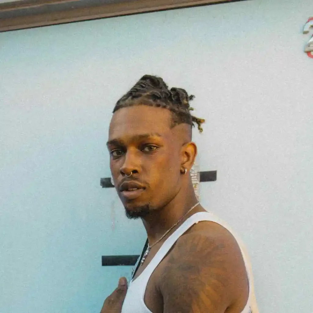 A man with braided hair showcasing long hair styling for men, posing confidently in a sleeveless white shirt against a light blue wall, decorated with a tattoo.