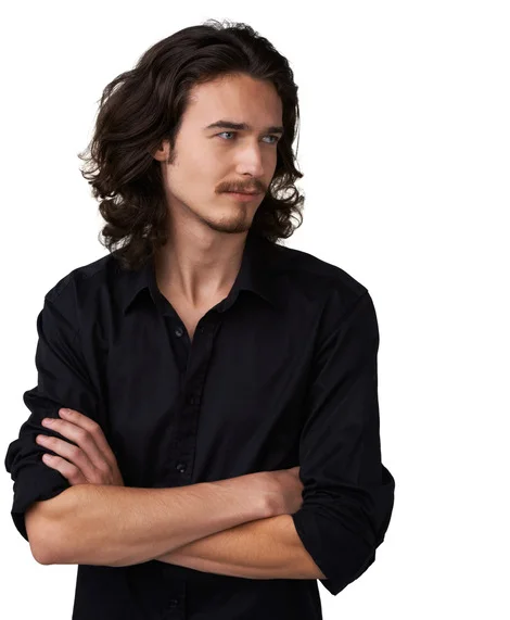 A young man showcasing one of the trendy summer hairstyles for men, with long, wavy hair, stands confidently with his arms crossed, wearing a black button-up shirt against a white background.