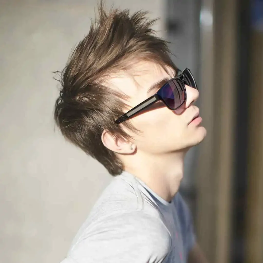 A person showcasing men hairstyles with glasses, featuring tousled hair, leaning back in a gray t-shirt against a softly blurred background.