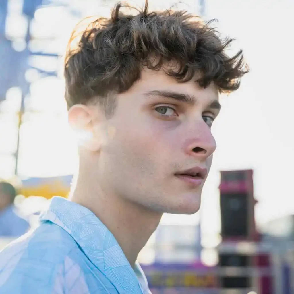 A person with men wavy hairstyles and curly hair, dressed in a light blue shirt, stands in a sunlit outdoor setting with colorful background elements.