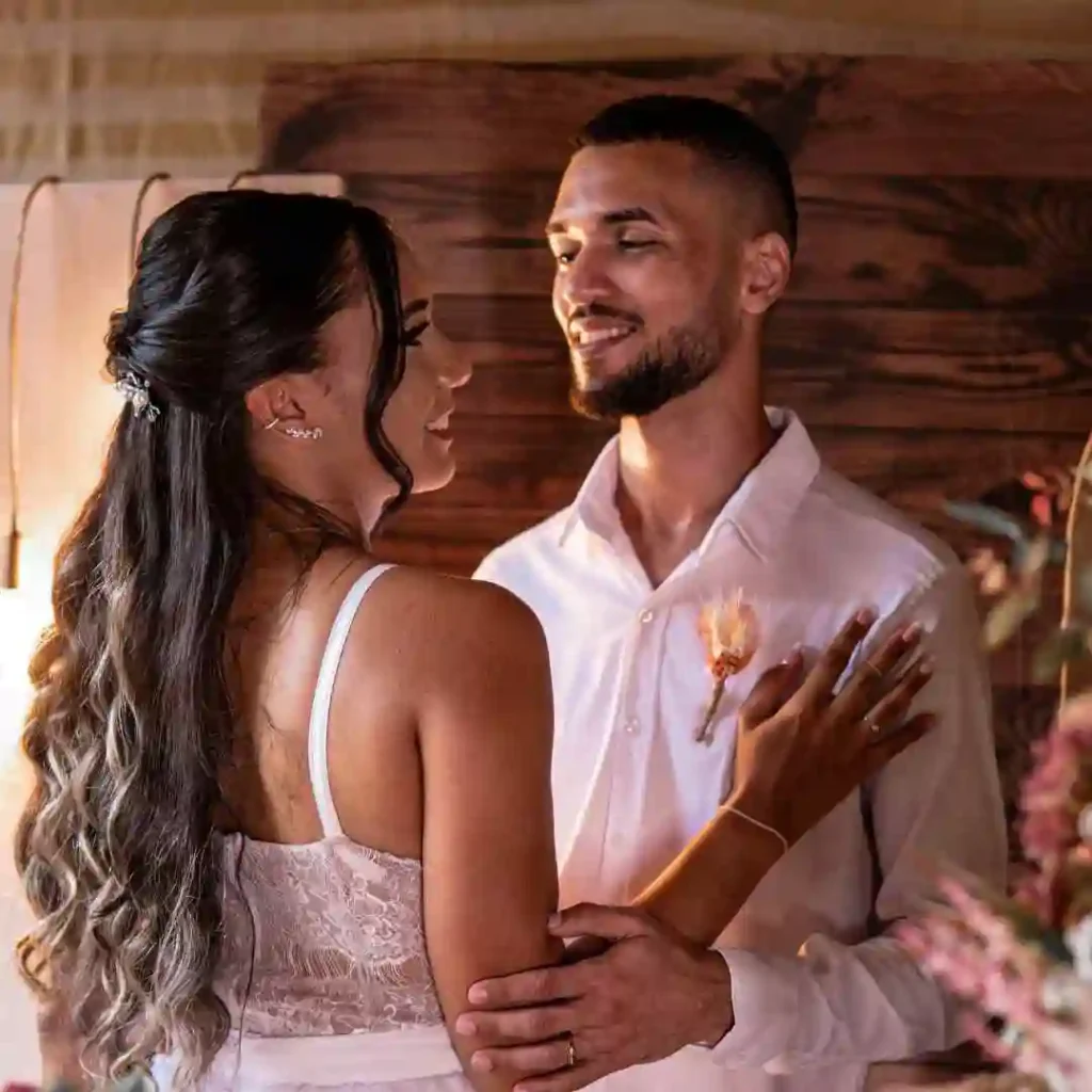 A couple shares an intimate moment, dressed elegantly; the woman in a lace dress, the man in a white shirt, showcasing a stylish wedding hairstyle for men, surrounded by warm decor.