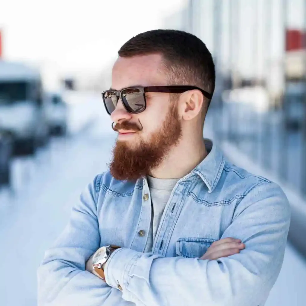 A person with a beard stands confidently with arms crossed, showcasing trendy men hairstyles with glasses, wearing a denim jacket and sunglasses against a blurred snowy backdrop.