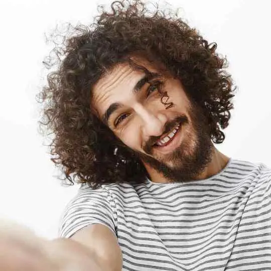 A person with men wavy hairstyles and curly hair wearing a striped shirt takes a selfie, extending their arm towards the camera against a white background.