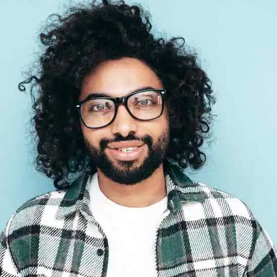 Person with curly hair showcasing one of the trendy men hairstyles with glasses, wearing a black and white checkered shirt over a white t-shirt, set against a pastel blue background.