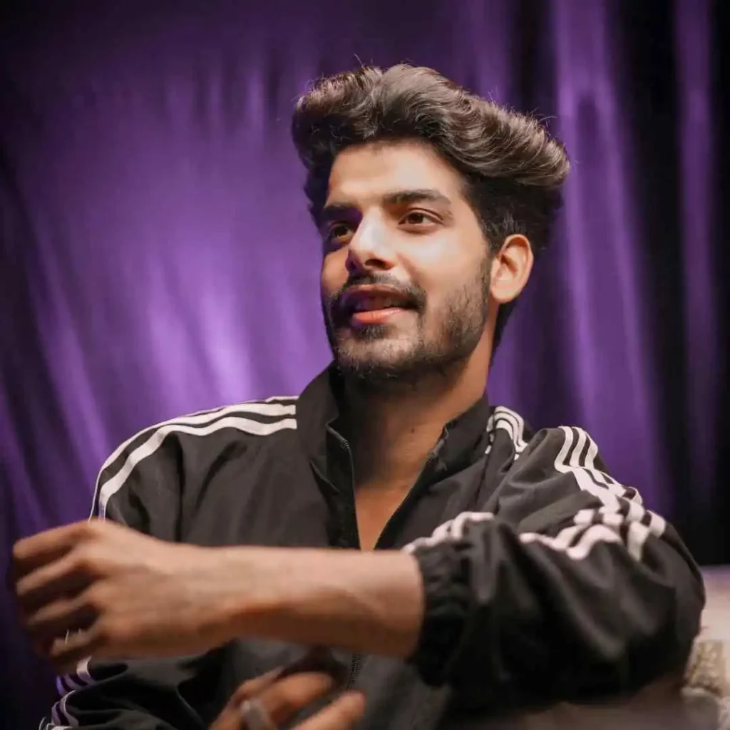 A person with men wavy hairstyles wearing a black jacket with white stripes rests their arm on a surface, against a backdrop of purple fabric.