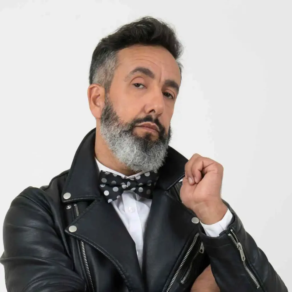 A stylish individual showcasing a Wedding Hairstyle For Men, wearing a leather jacket and polka dot bow tie, poses thoughtfully against a plain background.