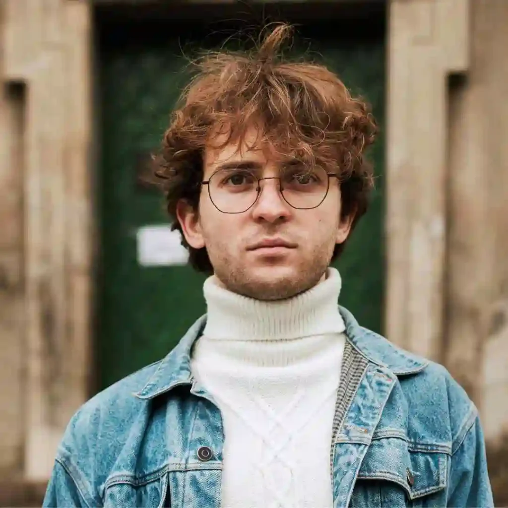 A person stands in front of a green door, wearing a denim jacket over a white turtleneck, with tousled brown hair and stylish glasses, showcasing trendy men hairstyles with glasses.