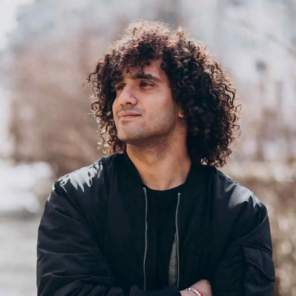 A person with curly hair showcasing long hair styling for men, posing in a black jacket with arms crossed against a blurred natural background.