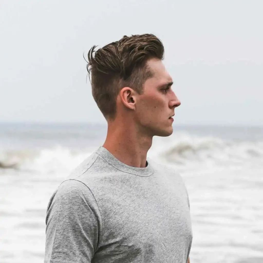 A person with men wavy hairstyles wearing a gray t-shirt stands by the ocean, gazing into the distance, with waves gently lapping at the shore.