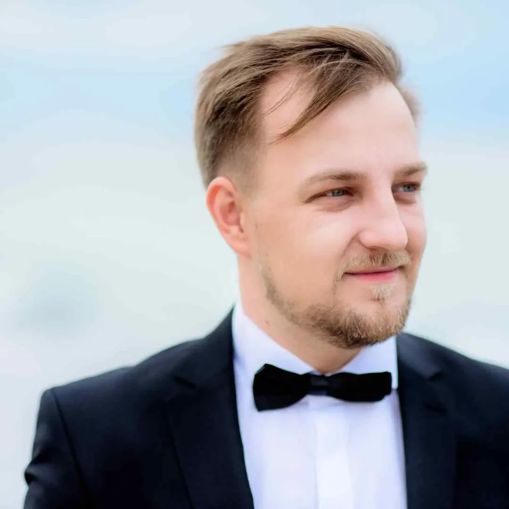 A man in a formal black tuxedo with a bow tie stands against a soft blue background, exuding elegance and sophistication, showcasing a classic wedding hairstyle for men.