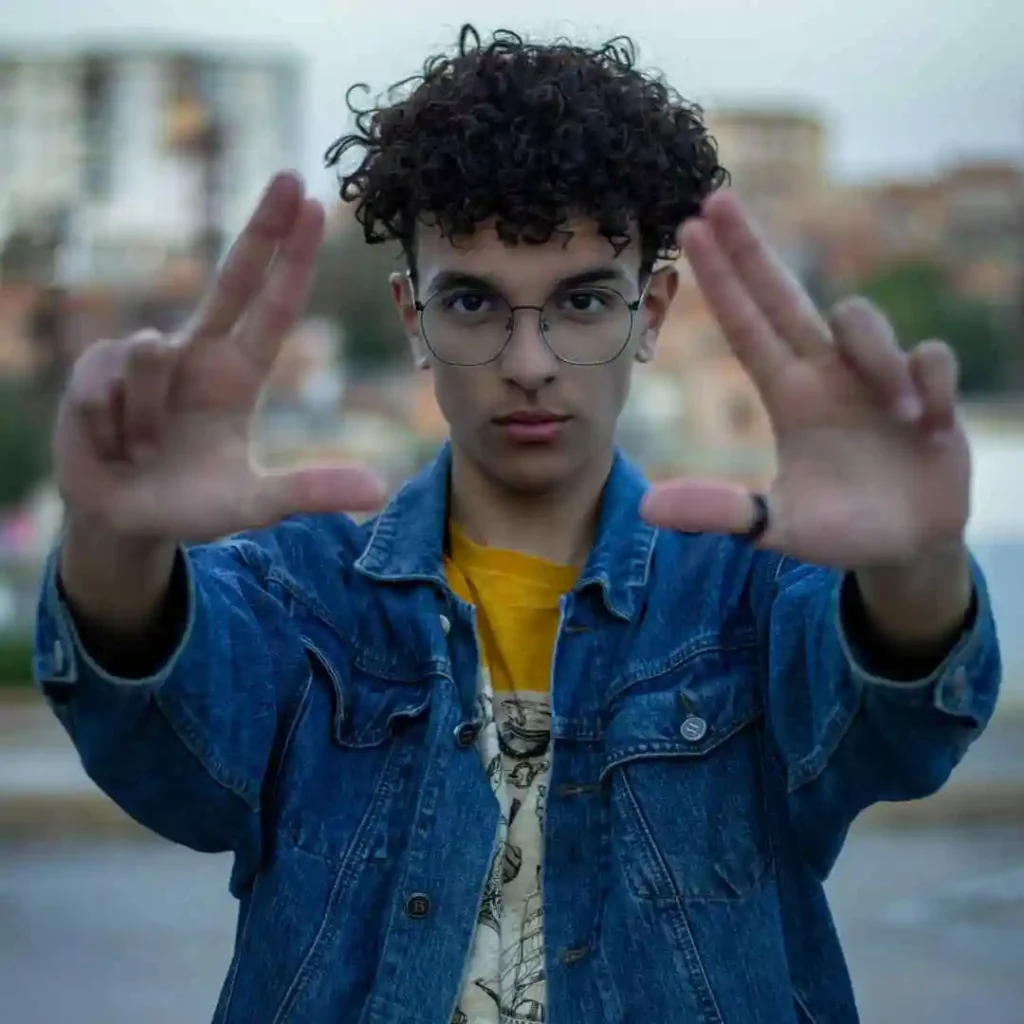 A man showcasing trendy men hairstyles with glasses, wearing a denim jacket and yellow shirt, holds his hands up in a framing gesture against a blurred urban background.