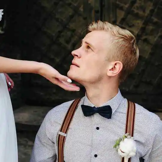 27 Wedding Hairstyles for Men That Can Elevate Your Style 1 A groom showcasing a stylish wedding hairstyle for men, dressed in a polka-dot shirt and bow tie, kneels and reaches for the hand of his bride in a flowing white gown.