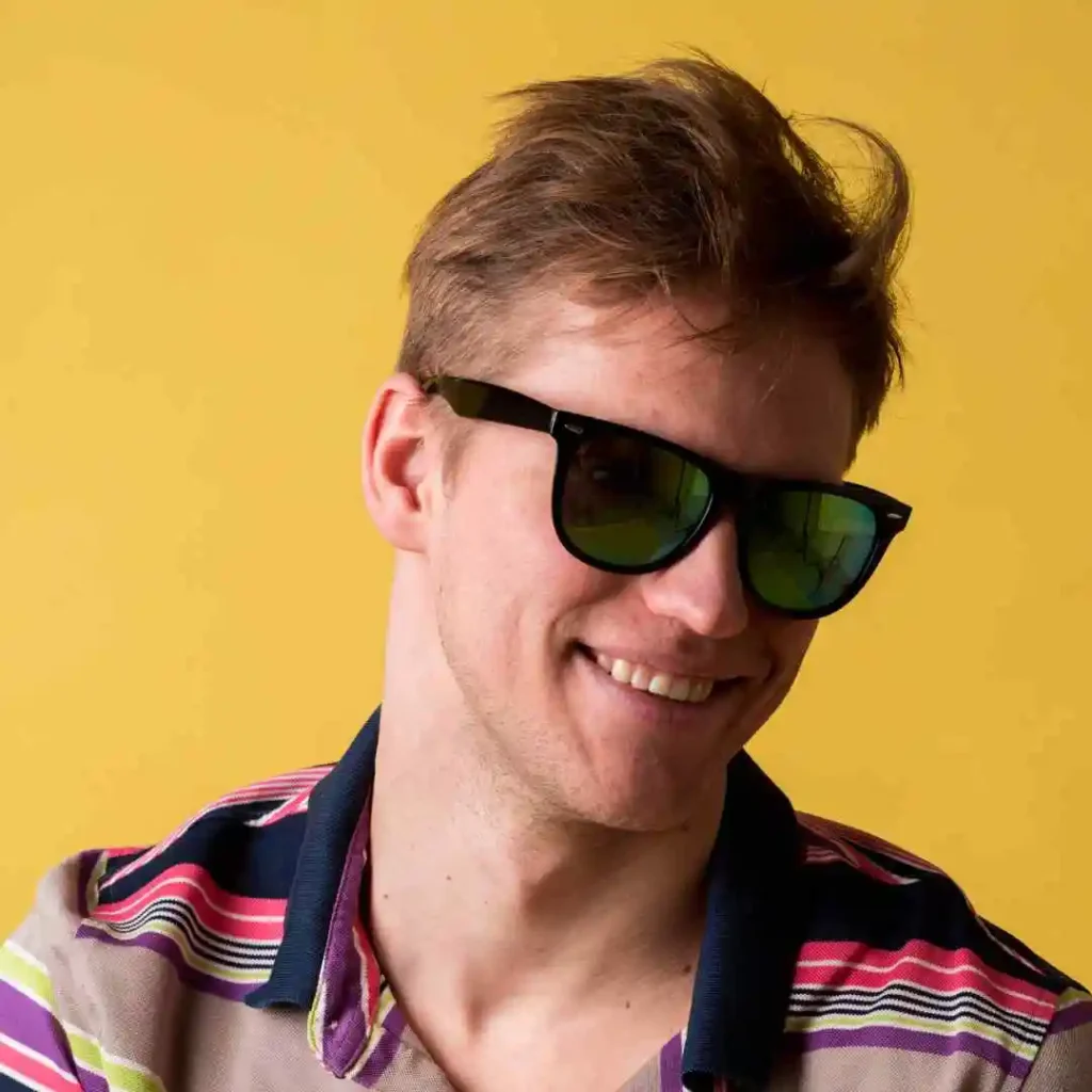 A young man with tousled hair showcasing men hairstyles with glasses, wearing a colorful striped shirt against a bright yellow background.