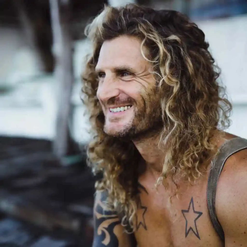 A man with shoulder-length curly hair styled in a trendy long hair styling for men, visible tattoos, wearing a tank top, posing in a rustic outdoor setting.