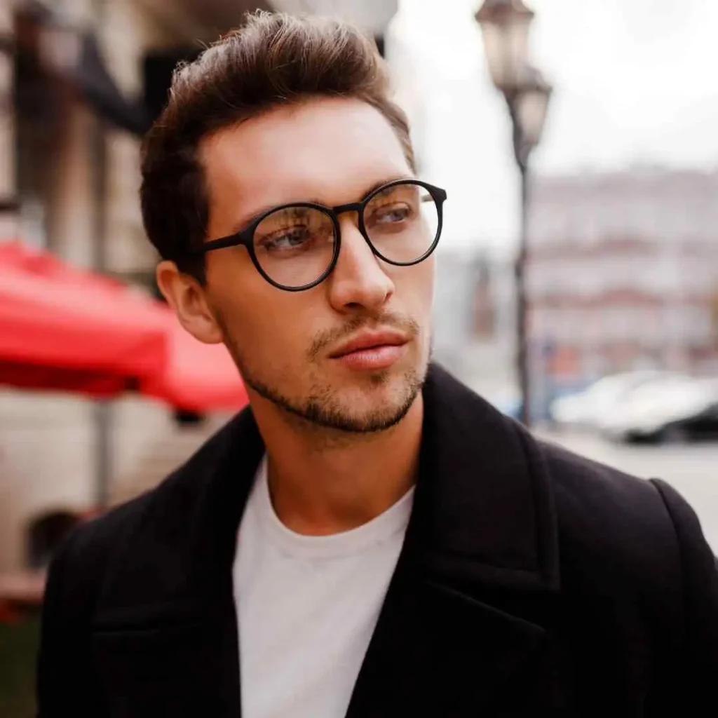A man showcasing men hairstyles with glasses, wearing a black coat over a white t-shirt, stands outdoors with urban scenery and a red umbrella in the background.