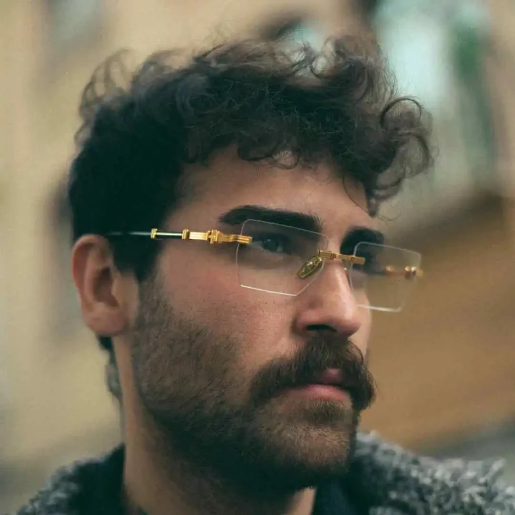 A person with curly hair and glasses showcasing trendy men hairstyles with glasses, standing outdoors, wearing a textured sweater, surrounded by a blurred, urban background.