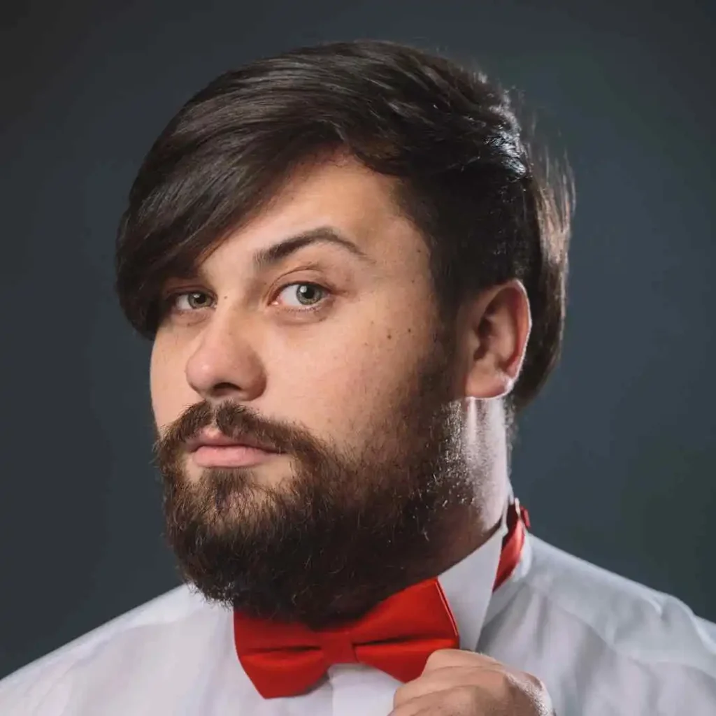 A man in a white shirt adjusts a red bow tie, set against a dark background, showcasing a sophisticated look and a stylish wedding hairstyle for men.